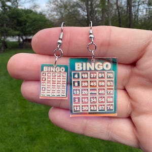 May include: A pair of novelty earrings featuring bingo cards. Each earring has a rectangular shape with a bingo card design, including numbers and the word "BINGO" at the top. The earrings are suspended from silver-colored fish hook ear wires.