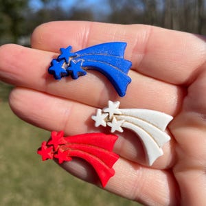 May include: Three plastic buttons shaped like shooting stars with white, red, and blue paint. Each button has three white stars.