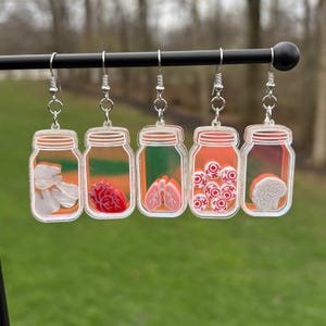 May include: Five pairs of novelty earrings. Each earring features a clear jar shape with a silver hook. The jars contain different anatomical elements: teeth, a heart, lungs, eyeballs, and a brain. The background of the jars is orange and green.