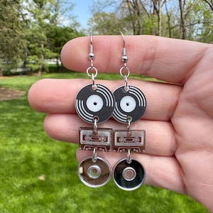 May include: Silver-tone dangle earrings featuring a retro music theme. Each earring has a black vinyl record design at the top, a cassette tape in the middle, and a clear CD at the bottom. The earrings are held up by silver hooks.