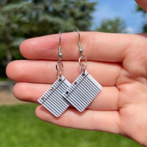 May include: A pair of dangle earrings featuring a white square design resembling a sheet of lined paper. The earrings have black lines and small holes along the left side, with silver-colored hooks and connecting rings. The earrings are held in a hand.