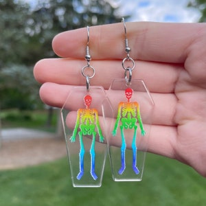 May include: A pair of rainbow skeleton earrings in clear coffin-shaped acrylic. The earrings are on silver hooks.