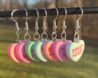 May include: Seven heart-shaped earrings in pastel shades of pink, green, and peach. Each earring has a silver hook and a different message in red text, including "SWEET HEART" and "HUG ME".