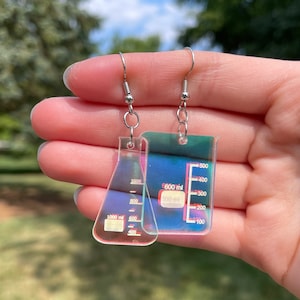 May include: A pair of iridescent earrings featuring a beaker and a graduated cylinder. The beaker has markings for 1000 ml, 900 ml, 800 ml, 700 ml, 600 ml, 500 ml, 400 ml, 300 ml, 200 ml, and 100 ml. The graduated cylinder has markings for 500 ml, 400 ml, 300 ml, 200 ml, and 100 ml.