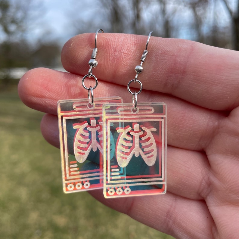 May include: A pair of silver dangle earrings with a clear acrylic charm featuring a stylized image of a chest x-ray with a red and white outline of lungs.