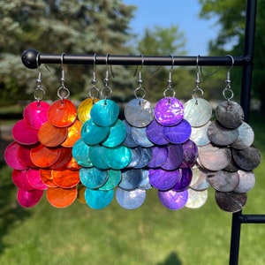 May include: Six pairs of iridescent shell earrings in a rainbow of colors, hanging from a black stand. The earrings are made of multiple layers of round shells, creating a cascading effect. The colors are pink, orange, teal, blue, purple, and gray.
