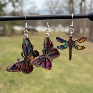 May include: Three wooden earrings: two butterfly designs and one dragonfly. The butterfly earrings have colorful stained-glass-like wings. The dragonfly earring has colorful wings. All earrings hang from silver-colored hooks.