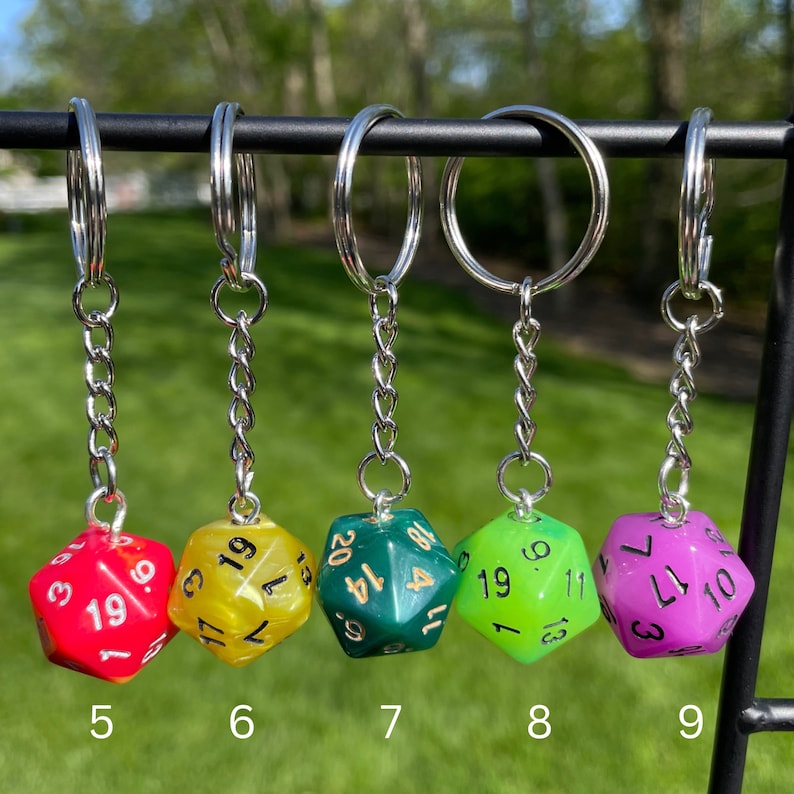 May include: Five keychains with different colored dice charms. The dice are numbered 1-20. The keychains are silver and have a circular ring at the top.