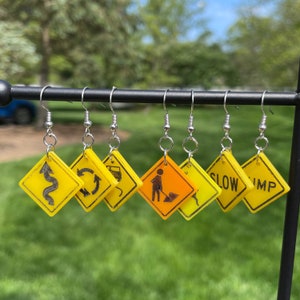 May include: A set of six yellow and orange diamond-shaped earrings with various road signs, including a winding road, a roundabout, a construction worker, and the words "Slow" and "Bump".
