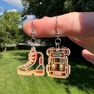 May include: A pair of dangle earrings featuring a hiking boot charm and a backpack charm. The charms are made of translucent orange and yellow acrylic with etched outlines, suspended from silver-toned metal fishhook ear wires.