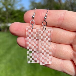 May include: Clear acrylic earrings with a rectangular shape and a white checkered pattern. The earrings have silver-colored fish hook ear wires. The background is a blurred green.