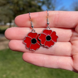 May include: A pair of silver earrings with red poppy flower charms. The flowers have black centres and are outlined in silver.