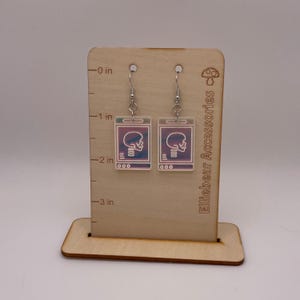 May include: A pair of iridescent earrings featuring a skull image on a white background. The earrings are hanging from silver hooks on a wooden display stand with a ruler.