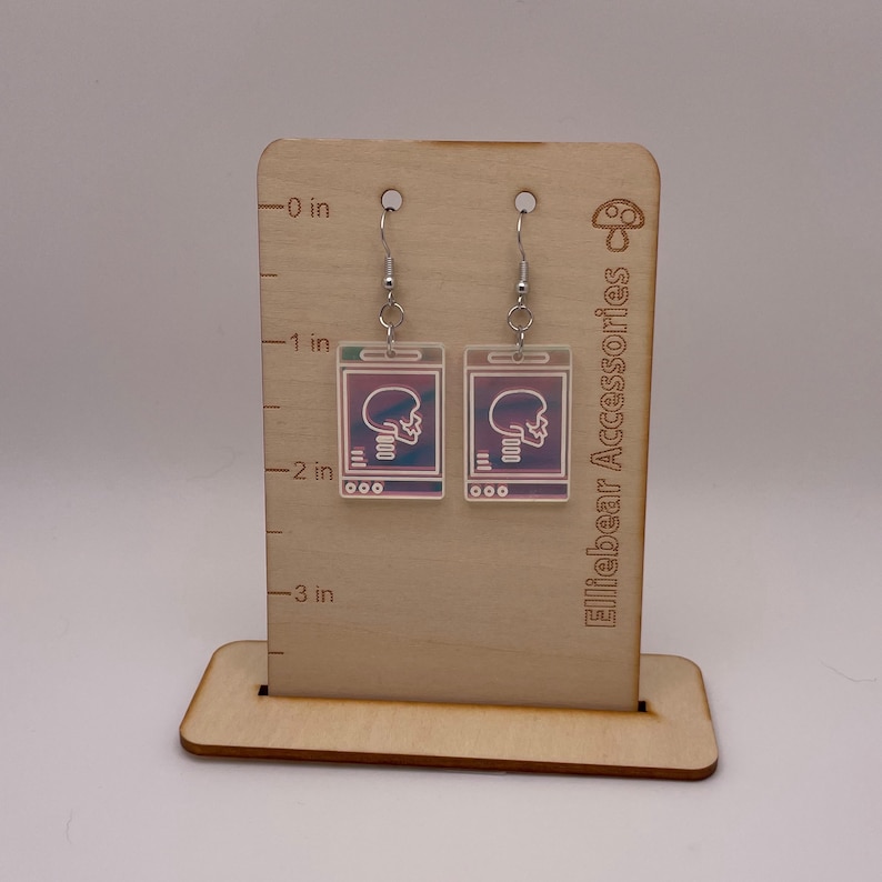 May include: A pair of iridescent earrings featuring a skull image on a white background. The earrings are hanging from silver hooks on a wooden display stand with a ruler.