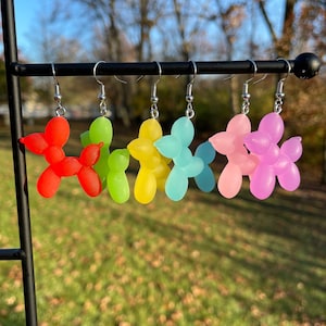 May include: Five pairs of balloon dog earrings in different colors. The earrings are made of a translucent plastic material and are shaped like balloon dogs. The colors are red, green, yellow, blue, and pink.