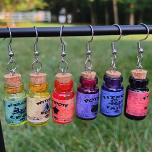 May include: Six potion bottle earrings with cork stoppers and silver-tone hooks. Each bottle is a different color, with labels reading "Frog Legs", "Witch Brew", "Antidote", "Potion", "Lizard Tail", and "Eye of Newt".