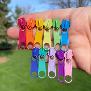 May include: A set of ten colorful zipper pulls in a rainbow pattern. The zipper pulls are small and made of metal. They are arranged in a row, with the red zipper pull at the top and the pink zipper pull at the bottom.