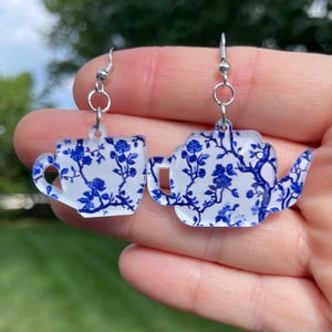May include: A pair of earrings featuring a teacup and teapot design. The earrings are white with a blue floral pattern and silver-colored hooks. The teacup and teapot are cut out shapes.