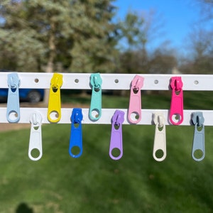 May include: Ten colorful zipper pulls hanging on a white rack. The zipper pulls are in the following colors: light blue, yellow, light blue, pink, hot pink, white, light purple, blue, and gray.