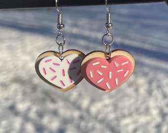 May include: A pair of heart-shaped earrings with pink and white frosting designs. The earrings are made of wood and have silver hooks.