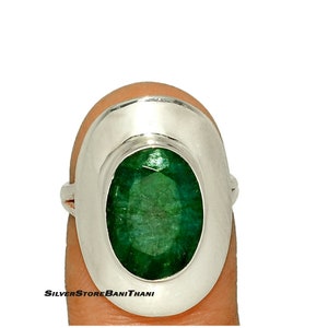 May include: A silver ring with a large, oval-shaped green emerald gemstone set in a bezel setting. The ring has a wide band and a smooth, polished finish.