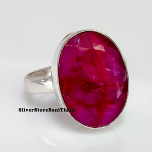 May include: A silver ring with a large, oval-shaped, magenta gemstone. The gemstone has a faceted surface. The ring band is polished silver. The jewellery is set against a white background. The ring is a statement piece.