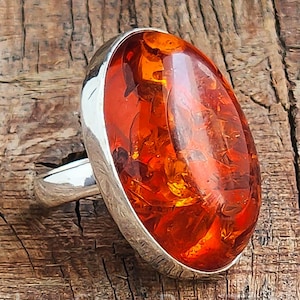 以下が含まれることがあります： 銀色のバンドが付いた、楕円形の琥珀の宝石リング。琥珀は、内部のインクルージョンを持つ、豊かで半透明なオレンジ色をしています。リングは木製の表面に置かれています。