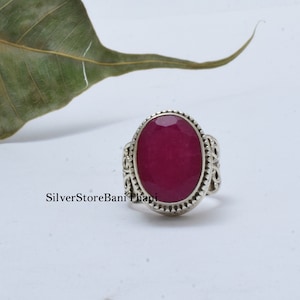May include: A silver ring featuring an oval-shaped, deep pink gemstone set in an ornate silver bezel. The ring has a detailed, intricate design.