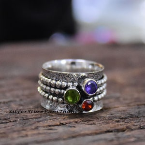 Könnte beinhalten: Ein drehbarer Silberring mit drei Edelsteinen: einem violetten Amethyst, einem grünen Peridot und einem roten Granat.
