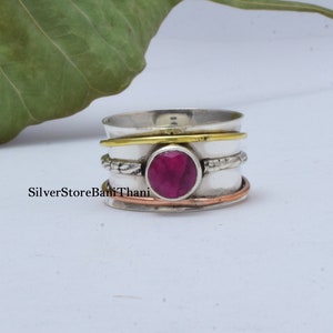 May include: A silver spinning ring with a red gemstone and three bands of different metals: silver, gold, and copper.
