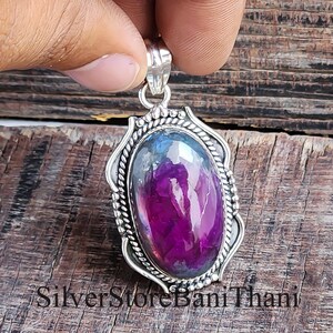 May include: A silver pendant featuring an oval, polished, purple gemstone. The gemstone displays a mix of purple and blue tones. The pendant has a decorative silver frame with a rope-like design. The text "Silver Store Bani Thani" is at the bottom.