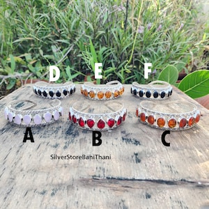 May include: Ten silver bracelets with different colored gemstones. The bracelets are arranged in a row on a wooden surface. The bracelets are labeled A through F. The text "SilverStoreBaniThani" is visible below the bracelets.