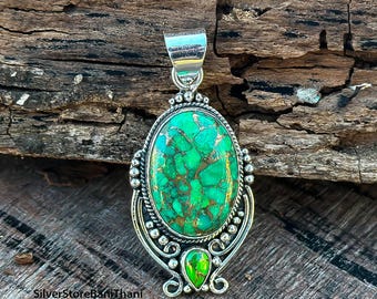 May include: Oval pendant with a vibrant green and gold stone set in a silver frame. The pendant has a detailed design with a bail at the top for a chain. The stone has a unique, textured appearance. The pendant is displayed on a wooden surface.