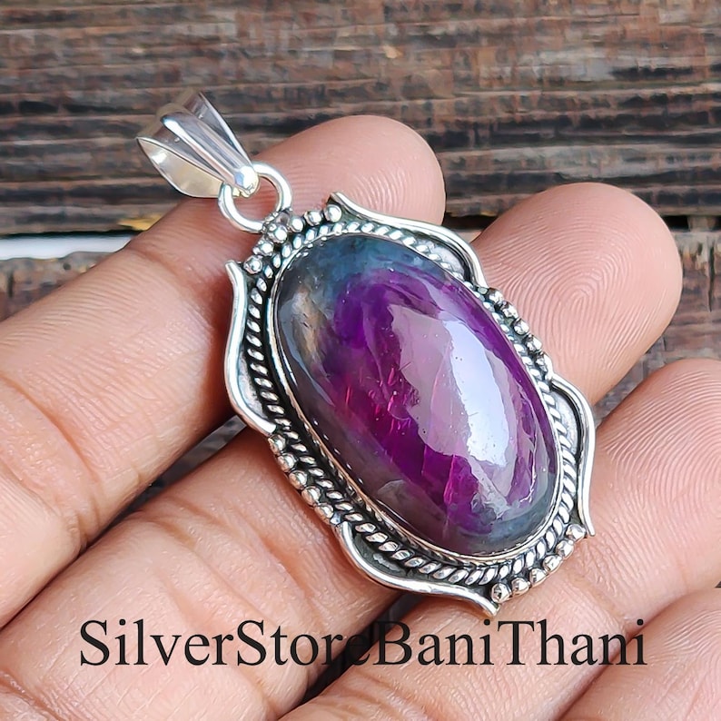 May include: Oval pendant featuring a large, polished purple gemstone set in a silver frame. The frame has a decorative rope-like border and a bail for a chain. The text "SilverStor Bani Thani" is at the bottom.
