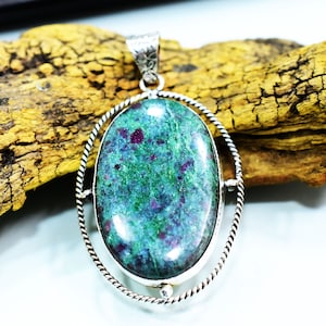 May include: A silver pendant featuring a green and red oval gemstone set within a twisted silver frame. The pendant is resting on a piece of driftwood.