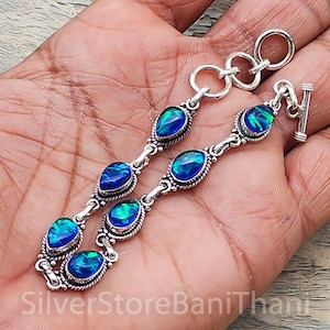 Op de afbeelding: Een zilveren armband met ovale en druppelvormige blauwe opaalstenen. De stenen zijn gezet in sierlijke zilveren zettingen en verbonden door delicate schakels. De armband heeft een tuimelsluiting.