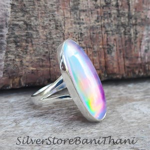 Op de afbeelding: Een zilveren ring met een grote, ovale iriserende edelsteen. De steen toont roze, paarse en gele tinten. De ring heeft een gespleten schachtontwerp. De tekst "SilverStoreBaniThani" is zichtbaar aan de onderkant.