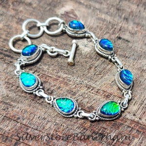 May include: A silver bracelet featuring teardrop-shaped, iridescent blue and green gemstones. The bracelet has a toggle clasp and is set on a wooden surface. The gemstones are framed by a silver rope-like design.