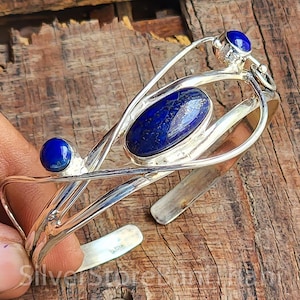 May include: A silver cuff bracelet featuring three blue lapis lazuli stones. The bracelet has an open design with an oval stone in the center and two round stones on the ends. The metal has a polished finish.