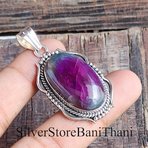 Peut inclure: Un pendentif en argent avec une pierre précieuse ovale, violet foncé. La pierre est sertie dans un cadre argenté décoratif avec une bordure en forme de corde. Le pendentif a une bélière en argent. Le texte "SilverStoreBaniThani" est en bas.