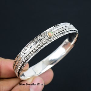 May include: Silver bangle bracelet featuring an opal gemstone and intricate engraved details. The bracelet is made of three bands with different textures.