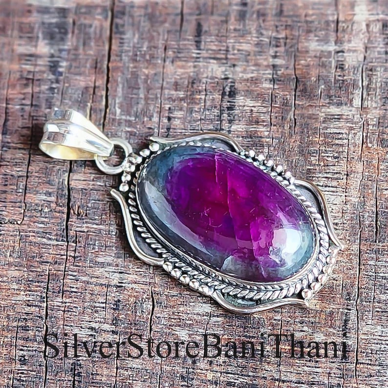 May include: A silver pendant featuring an oval, deep purple gemstone. The gemstone is set in a decorative silver frame with a rope-like border and small silver beads. The pendant has a silver bail for a chain. The text "Silver Store Bani Thani" is at the bottom.