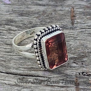 May include: A sterling silver ring with a rectangular, reddish-brown gemstone. The ring features a detailed, beaded border around the stone and a split shank design. The metal is stamped with "925".