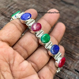 May include: A silver bracelet with a chain link design and oval-shaped gemstones in blue, red, and green.  The bracelet is on a hand.