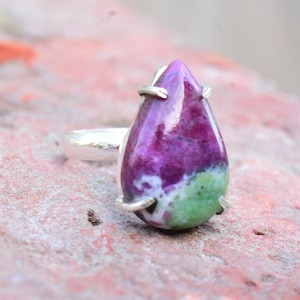 May include: A silver ring with a pear-shaped green, pink, and white gemstone. The gemstone is set in a silver bezel.