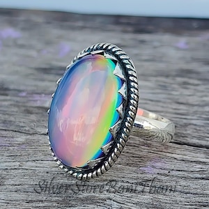 Könnte beinhalten: Silberner Ring mit einem ovalen, schillernden Stein. Der Stein zeigt ein Farbspektrum von Rosa, Blau und Gelb. Der Ring hat ein strukturiertes Band und einen dekorativen Rand mit Seil-Design und kleinen Zacken.