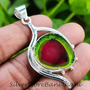 May include: A silver pendant with a watermelon tourmaline gemstone. The gemstone has a red centre, encircled by a green border, set in a decorative silver frame. The pendant has a loop for a chain.