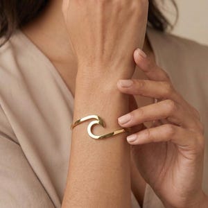 Può includere: Un bracciale rigido in metallo dorato lucido con un design a onda stilizzata. La fascia metallica presenta una superficie liscia e riflettente e una struttura aperta che si curva per formare un'onda che si infrange sul polso.