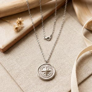 May include: A silver-toned metal necklace featuring a circular pendant with a raised four-pointed star design at the center, surrounded by intricate embossed patterns. The pendant hangs from a delicate silver-toned cable chain with a lobster clasp.