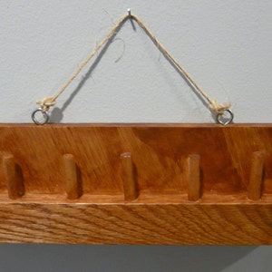 May include: A wooden wall-mounted key holder with six hooks. The key holder is stained a dark brown color and has a rope hanging from it.